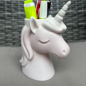 Könnte beinhalten: Ein hellrosa Einhorn-Schreibtisch-Organizer mit Stiften und Textmarkern. Das Einhorn hat ein spiralförmiges Horn, geschlossene Augen und eine fließende Mähne. Der Organizer besteht aus einem glatten Material.