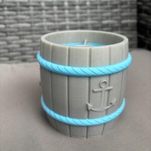 Könnte beinhalten: Ein grau-farbener, fassförmiger Behälter mit vertikalen Holzlatten und einem hellblauen Seil-Design oben und unten. Auf der Seite befindet sich ein Anker-Emblem. Dekoration im nautischen Stil.
