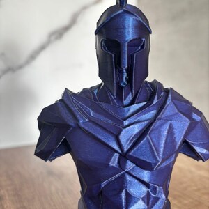 Könnte beinhalten: Dunkelblaue Büste eines spartanischen Kriegers mit Helm und hohem Kamm und detaillierter Rüstung. Die Skulptur hat ein geometrisches, facettiertes Design, das ihr eine moderne Ästhetik verleiht. Die Büste wird auf einer Holzoberfläche ausgestellt.