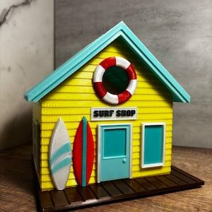 Op de afbeelding: Een miniatuur geel Surf Shop model met een turquoise dak en deur, en een bruin houten dek. De winkel heeft een rood-witte reddingsboei, twee surfplanken en de tekst "SURF SHOP" boven de deur.