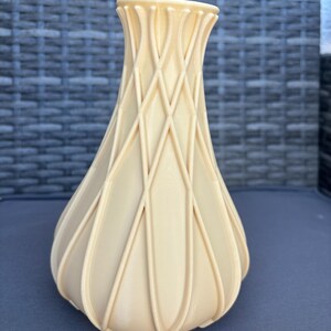 Könnte beinhalten: Eine beige dekorative Vase mit einem einzigartigen Gitterdesign. Die Vase hat einen bauchigen Boden und eine ausgestellte Oberseite. Das Äußere weist eine Reihe sich kreuzender Linien auf, die ein strukturiertes, geometrisches Muster bilden. Die Vase besteht aus einem festen, undurchsichtigen Material.