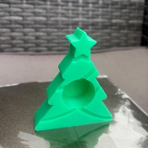 Könnte beinhalten: Ein hellgrüner Kerzenhalter in Form eines Weihnachtsbaums. Der Kerzenhalter hat einen Stern oben und eine kreisförmige Öffnung in der Mitte. Der Baum besteht aus einem festen Material und steht auf einer reflektierenden Oberfläche.