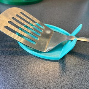 Könnte beinhalten: Eine türkisfarbene Kunststoff-Löffelablage hält einen Edelstahl-Pfannenwender. Die Löffelablage ist oval geformt und hat einen erhöhten Rand, um Tropfen aufzufangen. Der Pfannenwender hat ein geschlitztes Design.
