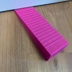 Könnte beinhalten: Ein leuchtend pinkfarbener Türstopper mit geriffelter Oberfläche, der verhindert, dass Türen vollständig geschlossen werden. Der rechteckige Keil besteht aus einem festen Material und liegt auf einem Holzboden.
