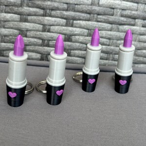 Könnte beinhalten: Vier Lippenstift-Schlüsselanhänger. Jeder Schlüsselanhänger hat einen weißen Körper, einen schwarzen Sockel mit einem rosa Herz und eine lila Lippenstiftspitze. Die Schlüsselanhänger sind auf einer grauen Oberfläche angeordnet.