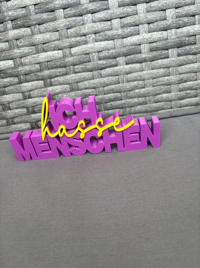 Op de afbeelding: Een paars 3D-geprint bord met de Duitse zin "Ich hasse Menschen". De woorden "Ich" en "Menschen" zijn in paarse blokletters, terwijl "hasse" in gele cursieve letters is geschreven.