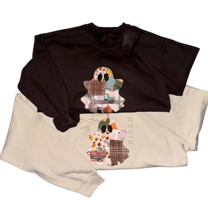 Pode incluir: São exibidas duas camisolas de manga comprida, uma castanha e outra creme. Cada uma apresenta um design de fantasma em patchwork com elementos temáticos de outono, como abóboras e um camião. A camisola creme tem o nome "PIPER" impresso verticalmente.