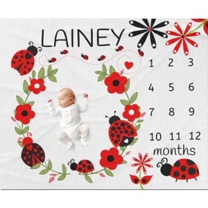 May include: A white baby blanket with a red and black ladybug theme. The blanket features a wreath of red flowers, green leaves, and ladybugs. The center of the blanket has a space for a baby to lay. The numbers 1 through 12 are printed on the blanket, along with the word "months". The name "Lainey" is printed at the top of the blanket.