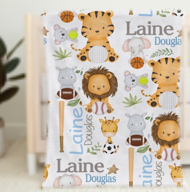 May include: White baby blanket featuring cartoon animals like tigers, lions, and elephants, along with sports equipment. The blanket includes the name "Laine Douglas" in blue lettering. The design is a mix of sports and safari themes.