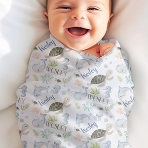 Op de afbeelding: Een witte baby-wikkeldeken met een blauw en groen oceaan-thema patroon. De deken heeft zeeschildpadden, walvissen, zeepaardjes en kwallen. De naam "Wesley" wordt herhaald in het hele patroon.