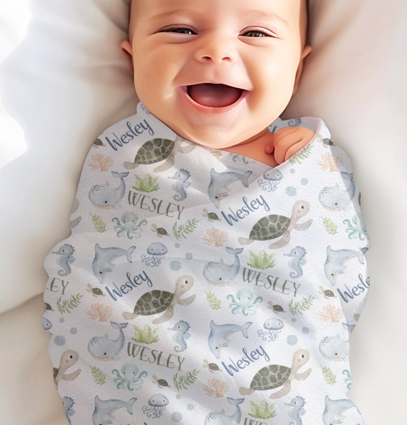 Op de afbeelding: Een witte baby-wikkeldeken met een blauw en groen oceaan-thema patroon. De deken heeft zeeschildpadden, walvissen, zeepaardjes en kwallen. De naam "Wesley" wordt herhaald in het hele patroon.