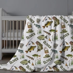 May include: White baby blanket with a repeating pattern of fish, fishing gear, and the name "OLIVER" in black. The blanket features illustrations of fish, fishing rods, hats, and tackle boxes. The blanket is draped over a crib.