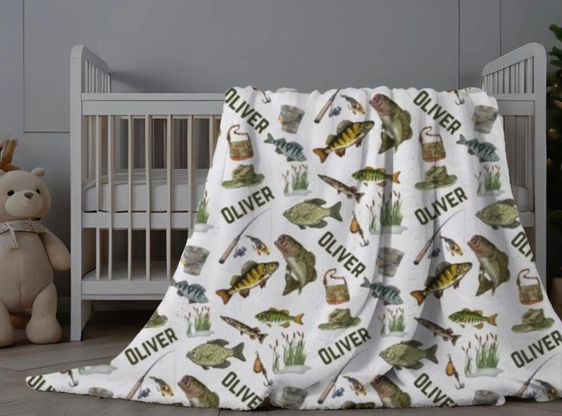 May include: White baby blanket with a repeating pattern of fish, fishing gear, and the name "OLIVER" in black. The blanket features illustrations of fish, fishing rods, hats, and tackle boxes. The blanket is draped over a crib.