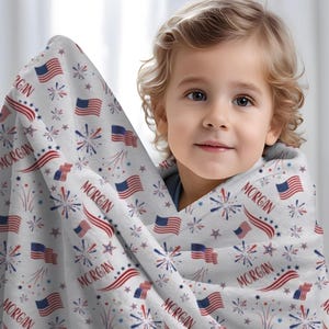 May include: A white baby blanket with a red, white, and blue American flag pattern. The blanket is decorated with stars, fireworks, and the name "Morgan".