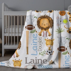 May include: A white baby blanket featuring a repeating pattern of cartoon animals, including a lion, giraffe, and tiger, with sports equipment. The blanket has the name "Laine Douglas" in blue and gray text.