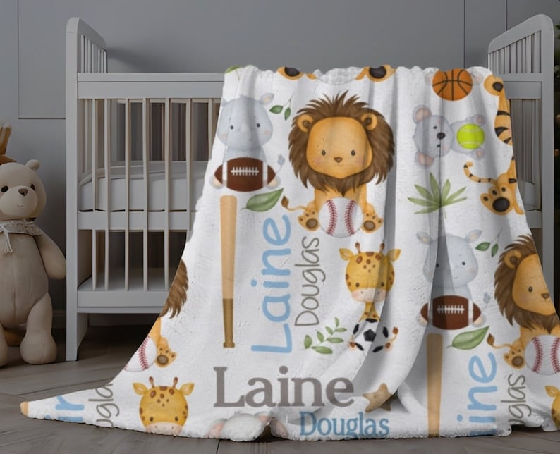 May include: A white baby blanket featuring a repeating pattern of cartoon animals, including a lion, giraffe, and tiger, with sports equipment. The blanket has the name "Laine Douglas" in blue and gray text.