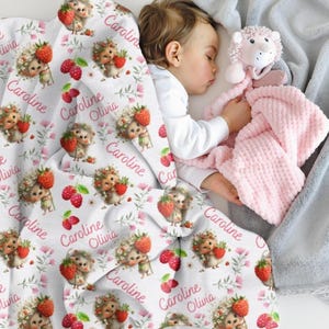 May include: A white fleece blanket featuring a repeating pattern of illustrated hedgehogs holding red raspberries, pink flowers, and the name Caroline Olivia in pink script. The blanket is draped over a sleeping child holding a pink plush toy.