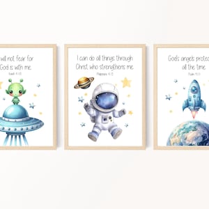 Op de afbeelding: Drie ingelijste prints met een ruimtethema. De eerste print toont een groene alien in een blauw ruimteschip met de tekst "Ik zal niet vrezen, want God is met mij. Jesaja 41:10". De tweede print toont een astronaut in een blauw ruimtepak met de tekst "Ik kan alles door Christus, die mij kracht geeft. Filippenzen 4:13". De derde print toont een blauwe raket die over een blauwe planeet vliegt met de tekst "Gods engelen beschermen mij altijd. Psalm 91:11".