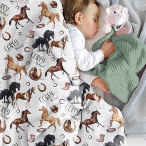 Pu&ograve; includere: Una coperta bianca per bambini con un motivo ripetuto di cavalli di vari colori, ferri di cavallo, stivali da cowboy e cappelli. Il nome "Corey Michael" &egrave; stampato sulla coperta. Un bambino che dorme &egrave; parzialmente visibile, insieme a una coperta verde e un peluche rosa.