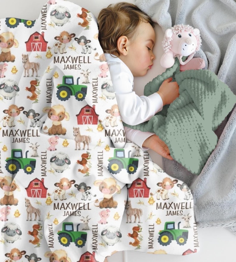 May include: A baby sleeps under a white blanket patterned with farm animals, tractors, and red barns. The name "MAXWELL JAMES" is printed on the blanket. A green textured blanket and a pink lion-shaped toy are also visible.