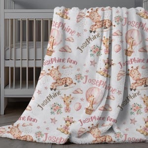 May include: A white plush blanket with a pink and brown giraffe print. The blanket features a repeating pattern of giraffes, hot air balloons, stars, and flowers. The name "Josephine" is repeated throughout the blanket.