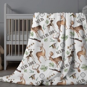 May include: White baby blanket with a hunting theme. The blanket features deer, dogs, rifles, and the name "Nolan James" in a repeating pattern. The blanket is draped over a white crib.