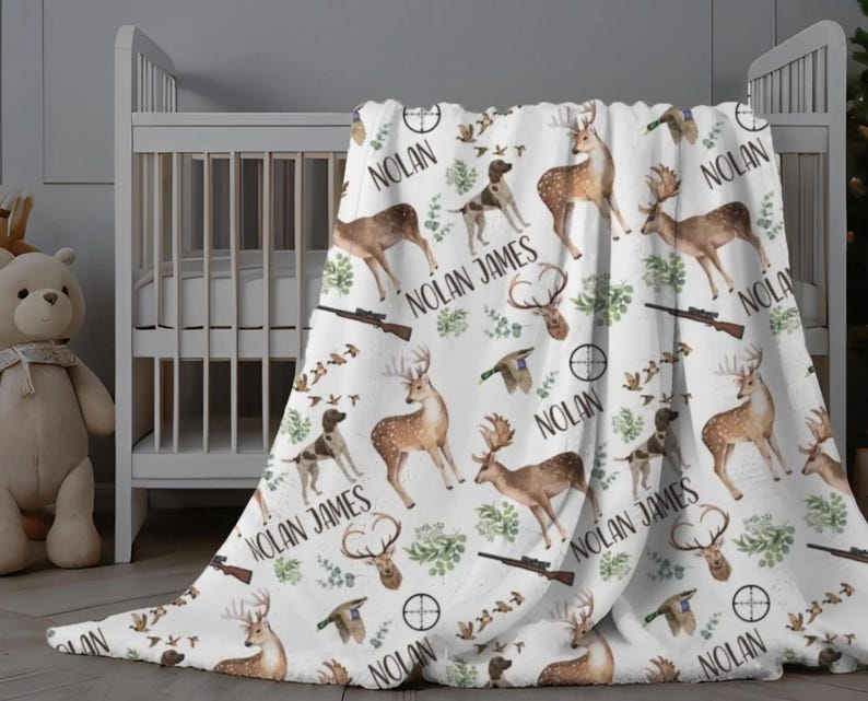 May include: White baby blanket with a hunting theme. The blanket features deer, dogs, rifles, and the name "Nolan James" in a repeating pattern. The blanket is draped over a white crib.