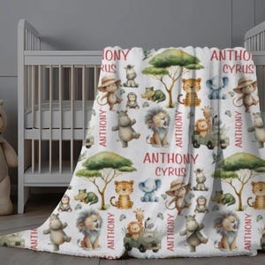 May include: A white baby blanket with a safari animal theme. The blanket features illustrations of lions, tigers, elephants, monkeys, and giraffes. The name "ANTHONY CYRUS" is printed in red. The blanket is draped over a white crib.