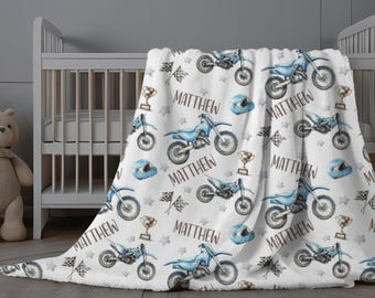 May include: White baby blanket with a repeating pattern of blue dirt bikes, helmets, trophies, and checkered flags. The name "MATTHEW" is printed throughout the design. The blanket is draped over a crib.
