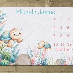 May include: A white baby milestone blanket with a blue and pink watercolor design featuring two sea turtles and coral. The blanket has the name "Mikaela Janine" and numbers 1-12 for tracking a baby's growth.