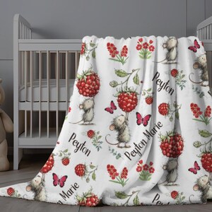 May include: White baby blanket with a whimsical design featuring cartoon mice, red raspberries, pink butterflies, and floral accents. The name "Peyton Marie" is printed on the blanket in a script font. The blanket is draped over a crib.