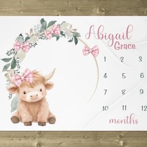 May include: A white milestone blanket featuring a watercolor illustration of a Highland cow with a pink bow. The blanket includes the name "Abigail Grace", a floral arch, and numbers 1-12 for monthly photos.