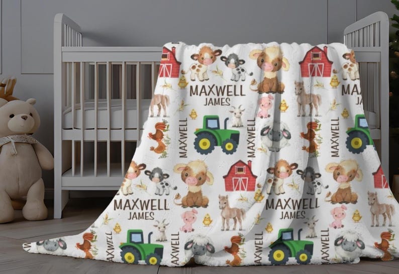 May include: White baby blanket with a farm animal pattern, including cows, pigs, and sheep. The blanket features a green tractor, red barn, and the name "MAXWELL JAMES" in black text. The blanket is draped over a white crib.
