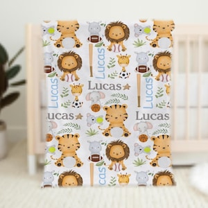 May include: A white baby blanket featuring a repeating pattern of cartoon animals, including tigers, lions, giraffes, and elephants. The blanket also includes sports equipment like baseball bats, footballs, and basketballs, with the name "Lucas" in blue.