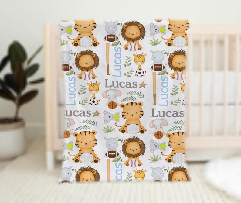 May include: A white baby blanket featuring a repeating pattern of cartoon animals, including tigers, lions, giraffes, and elephants. The blanket also includes sports equipment like baseball bats, footballs, and basketballs, with the name "Lucas" in blue.