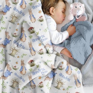 May include: A white baby blanket featuring a repeating pattern of a rabbit in a blue jacket, mushrooms, trees, and the name "Hendrix Dean" in blue script. A sleeping baby is partially covered by the blanket, with a blue plush toy.