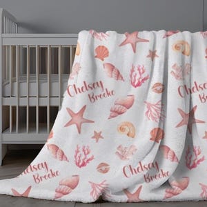 May include: A white fleece blanket with a pink and white seashell and starfish pattern. The blanket has the name "Chelsea Brecke" printed on it.