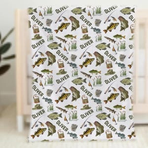 May include: White baby blanket with a repeating pattern of fish, fishing rods, hats, and the name "OLIVER" in black. The blanket features illustrations of various fish species and fishing-related items.