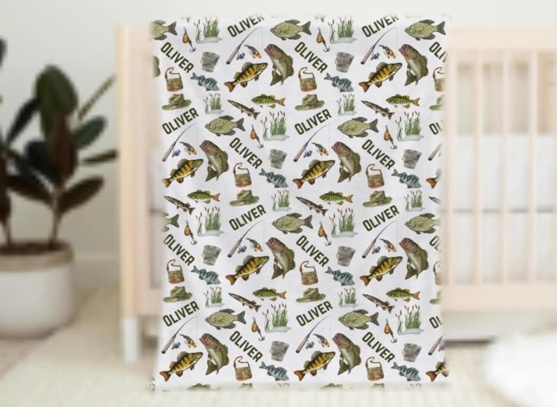 May include: White baby blanket with a repeating pattern of fish, fishing rods, hats, and the name "OLIVER" in black. The blanket features illustrations of various fish species and fishing-related items.
