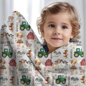 May include: A white blanket with a farm animal pattern, including cows, sheep, pigs, horses, and tractors. The name "MAXWELL JAMES" is printed repeatedly on the blanket. The blanket is wrapped around a child.