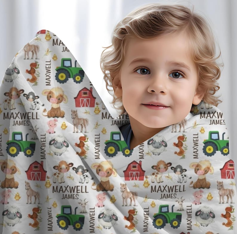 May include: A white blanket with a farm animal pattern, including cows, sheep, pigs, horses, and tractors. The name "MAXWELL JAMES" is printed repeatedly on the blanket. The blanket is wrapped around a child.