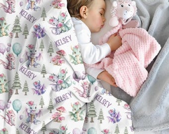 May include: A sleeping child is nestled under a white blanket adorned with whimsical watercolor dragons, trees, and balloons. The blanket is personalized with the name "Kelsey" in a soft purple font. A pink plush toy and a pink textured blanket are also present.