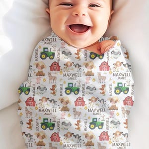 May include: A baby wrapped in a white swaddle blanket with a farm animal pattern. The pattern includes green tractors, red barns, cows, sheep, and horses. The name "MAXWELL JAMES" is printed throughout the design.