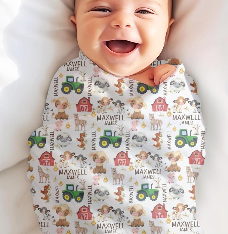 May include: A baby wrapped in a white swaddle blanket with a farm animal pattern. The pattern includes green tractors, red barns, cows, sheep, and horses. The name "MAXWELL JAMES" is printed throughout the design.