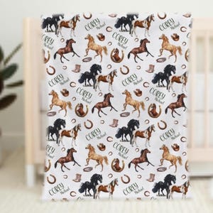 Pu&ograve; includere: Coperta per bambini bianca con un motivo ripetuto di cavalli di vari colori, ferri di cavallo, stivali da cowboy e il nome "Corey Michael". La coperta &egrave; drappeggiata su una culla.