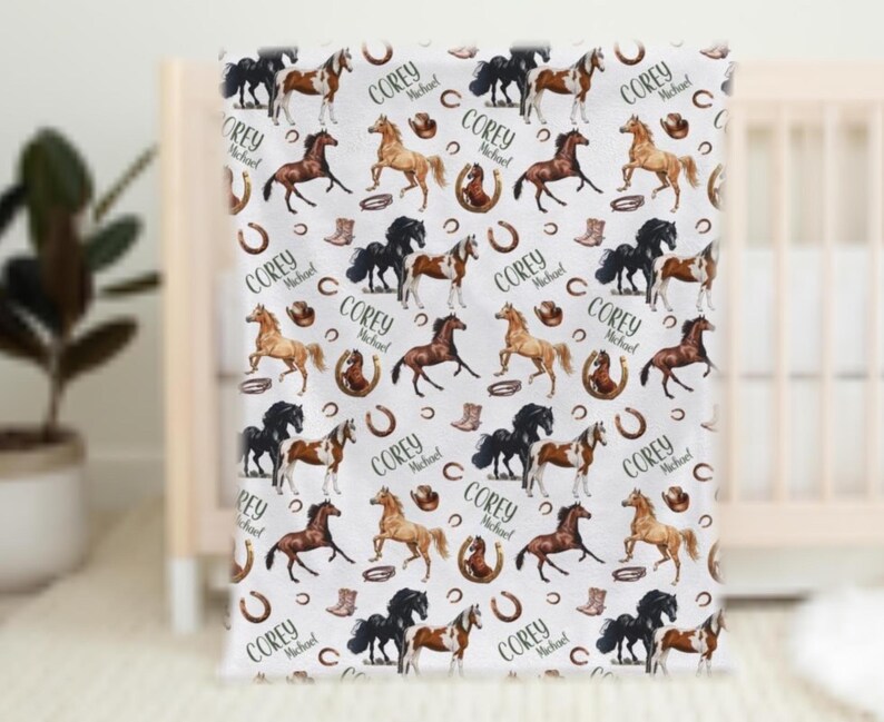Pu&ograve; includere: Coperta per bambini bianca con un motivo ripetuto di cavalli di vari colori, ferri di cavallo, stivali da cowboy e il nome "Corey Michael". La coperta &egrave; drappeggiata su una culla.
