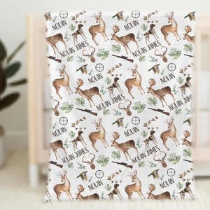 May include: White baby blanket with a repeating pattern of deer, hunting dogs, rifles, and foliage. The name "NOLAN JAMES" and "NOLAN" are printed throughout the design. The blanket is draped over a crib.