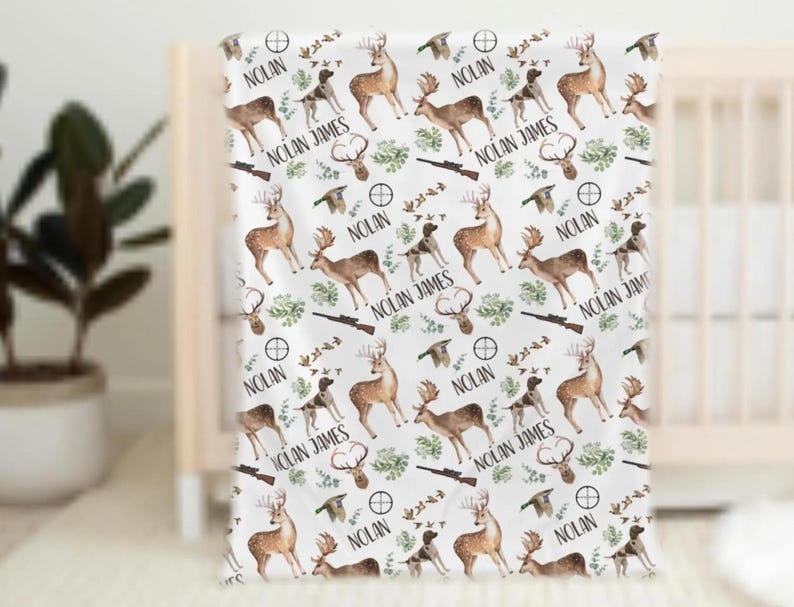 May include: White baby blanket with a repeating pattern of deer, hunting dogs, rifles, and foliage. The name "NOLAN JAMES" and "NOLAN" are printed throughout the design. The blanket is draped over a crib.