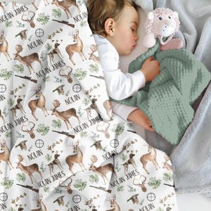 May include: A white baby blanket with a hunting theme, featuring deer, dogs, rifles, and the name "NOLAN JAMES" in black. A sage green textured blanket and a light pink lion toy are also visible.