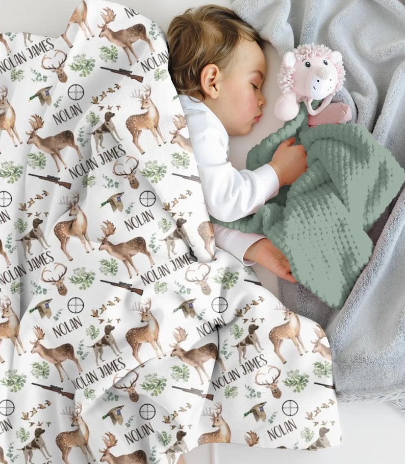 May include: A white baby blanket with a hunting theme, featuring deer, dogs, rifles, and the name "NOLAN JAMES" in black. A sage green textured blanket and a light pink lion toy are also visible.